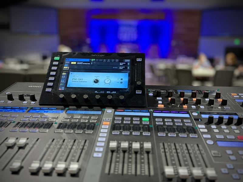 Close-up of a digital mixing console with a touchscreen and faders, with a blue-lit stage softly out of focus behind