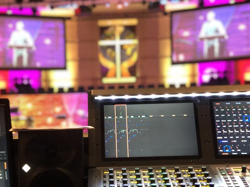 Front-of-house console and screens facing a sanctuary with a lit cross and a large screen showing a speaker at a podium, with colored wash lighting on the walls