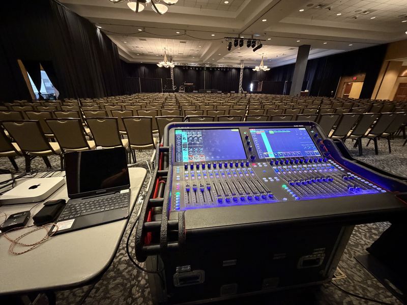 Digital mixing console with laptop set up at front of house in an empty ballroom with rows of chairs