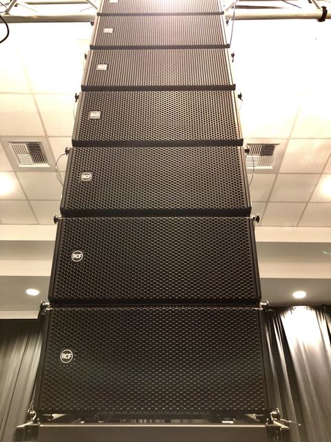 Looking straight up a flown line array of RCF speaker cabinets hanging from its frame under a ballroom ceiling