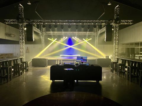 Empty venue with a truss lighting rig sending crossing yellow beams and a blue center beam over the stage, control table in the foreground