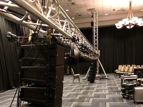 Line-array speaker cabinets flown from a truss bumper in a ballroom, with moving-head fixtures along the truss