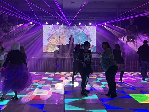 LED video wall behind a DJ at a costume party, with purple lasers and beam lights on truss overhead and dancers on a multicolor light-up dance floor